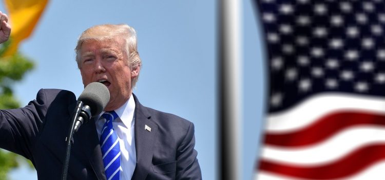 Photo of President Trump speaking at a microphone with one hand raised and an American flag behind him.