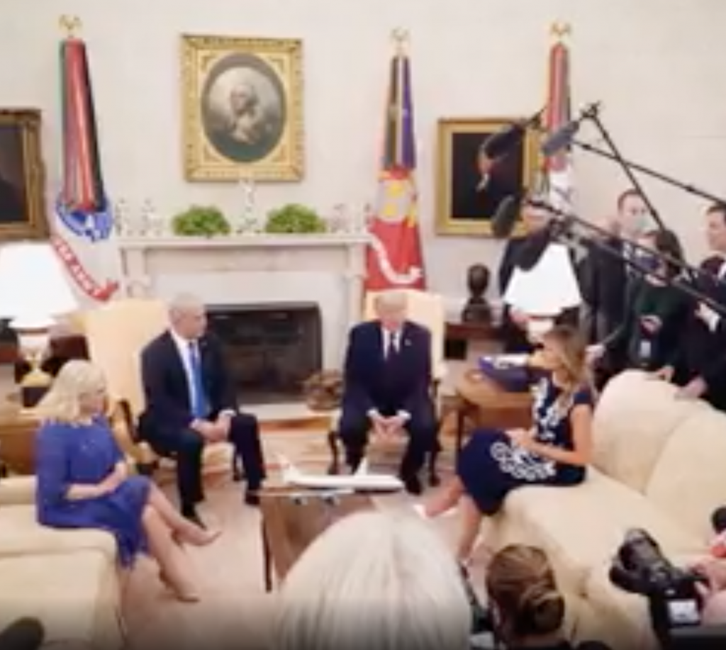 Paused video from a Trump ad. The scene shows President Trump and three other individuals sitting in the oval office with members of the press holding cameras and microphones to document the event.